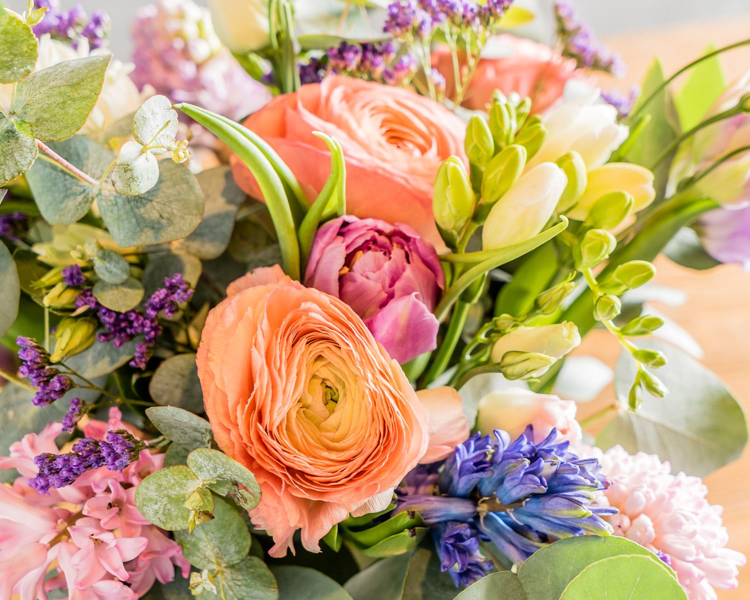 Close-up of a vibrant spring floral bouquet featuring peach ranunculus, purple hyacinths, and eucalyptus. Bright pastel petals and lush greenery evoke Easter and early spring.