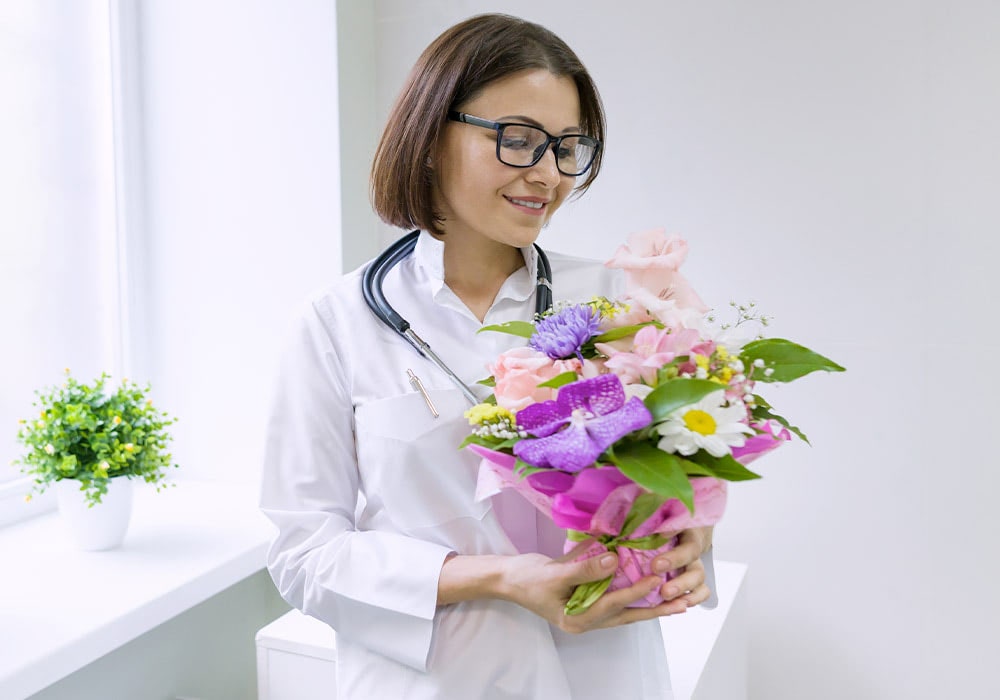 A nurse smiling while holding a colorful flower bouquet in a medical office, symbolizing National Nurses Week.