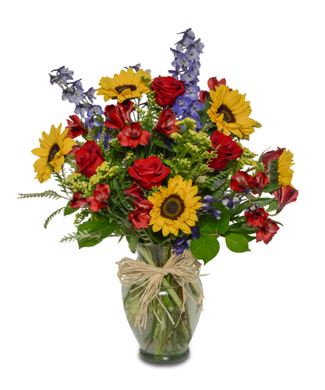 Vibrant bouquet of sunflowers, blue delphinium, red roses, and spray chrysanthemums in a clear glass vase.