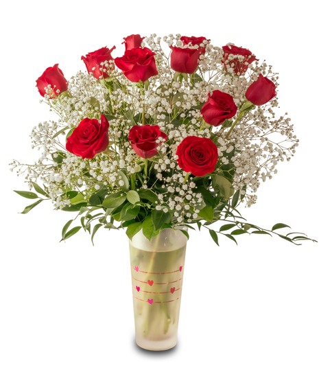 A romantic Valentine’s arrangement of fresh red roses and white baby’s breath arranged in a clear glass vase with heart accents and lush green foliage.