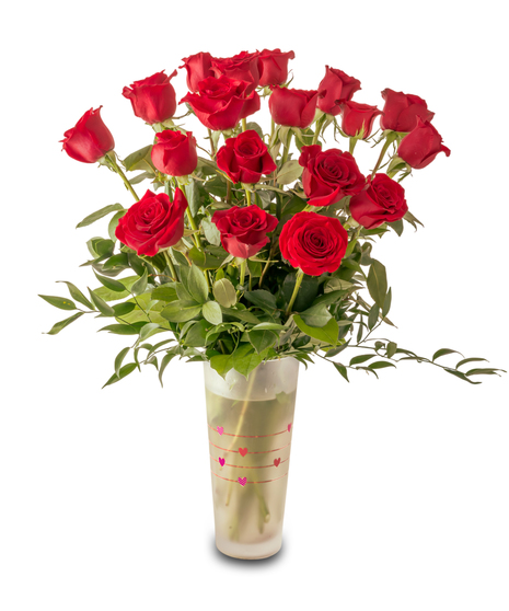 A romantic Valentine’s arrangement of fresh red roses and white baby’s breath arranged in a clear glass vase with heart accents and lush green foliage.