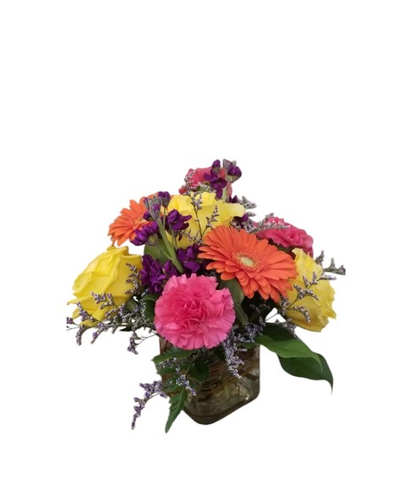 Bright floral arrangement with hot pink carnations, orange gerbera daisies, yellow roses, and purple stock in a glass cube vase.