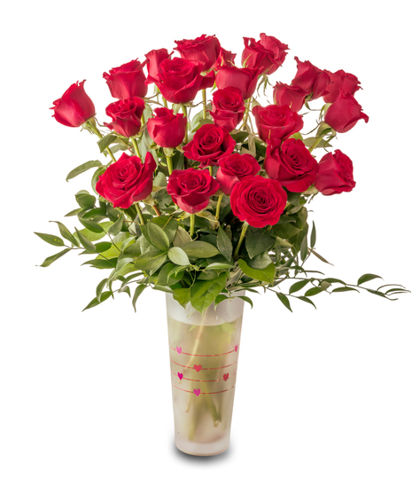 A romantic Valentine’s arrangement of fresh red roses and white baby’s breath arranged in a clear glass vase with heart accents and lush green foliage.