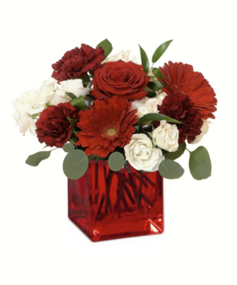 Red and white flower arrangement with roses, carnations, and gerbera daisies in a bold red glass cube vase, accented with greenery.