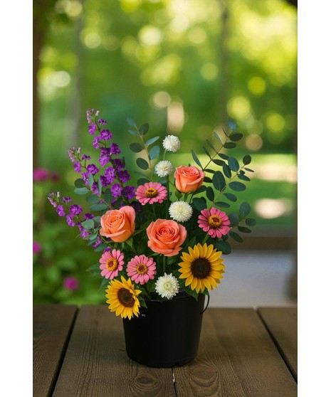 A black plastic bucket filled with assorted fresh flowers, including sunflowers, roses, daisies, larkspur, and eucalyptus, arranged upright against a simple dark backdrop.