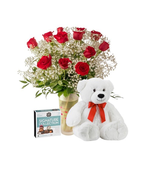 A romantic Valentine’s arrangement of fresh red roses and white baby’s breath arranged in a clear glass vase with heart accents and lush green foliage.