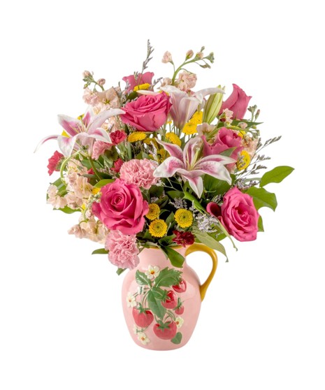 Pink roses, fragrant lilies, stock, and yellow accent flowers arranged in a pink strawberry pitcher vase, creating a cheerful pink and yellow bouquet with lush greenery.