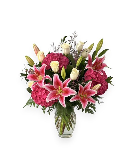 A glass vase filled with pink or blue hydrangeas, white roses, and fragrant lilies arranged with lush greenery, symbolizing joy and new beginnings.
