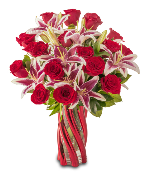 A floral arrangement featuring pink lilies with bright pink centers, red roses, and green foliage arranged in a red glass vase against a white background.