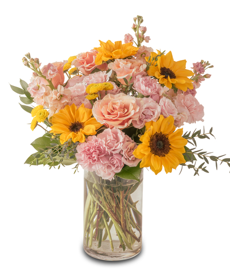 A clear glass vase filled with bright yellow sunflowers accented by soft pink and peach blooms, arranged with fresh greenery for a cheerful, sunny floral display.