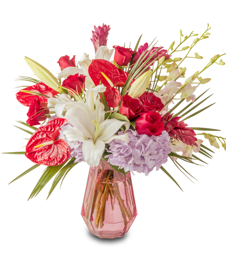 A tropical floral arrangement featuring red anthuriums, white lilies, red roses, and lavender hydrangeas arranged in a pink glass vase with greenery.