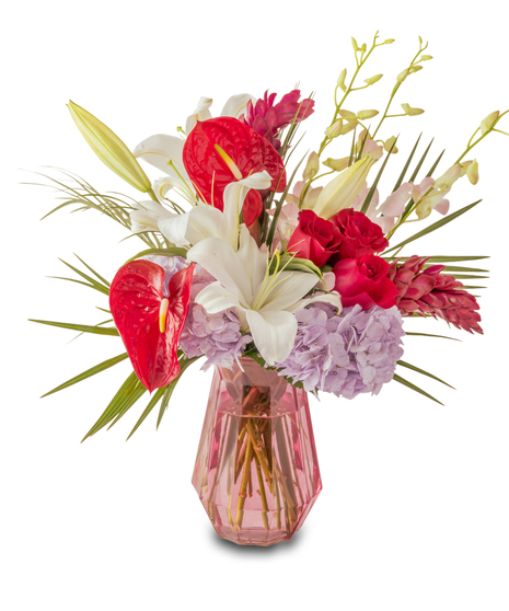 A tropical floral arrangement featuring red anthuriums, white lilies, red roses, and lavender hydrangeas arranged in a pink glass vase with greenery.